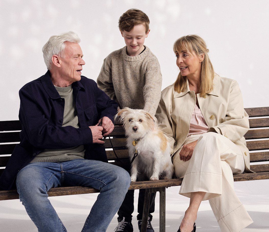 Middle aged couple and dog sitting on bench with a young boy standing and petting the dog.