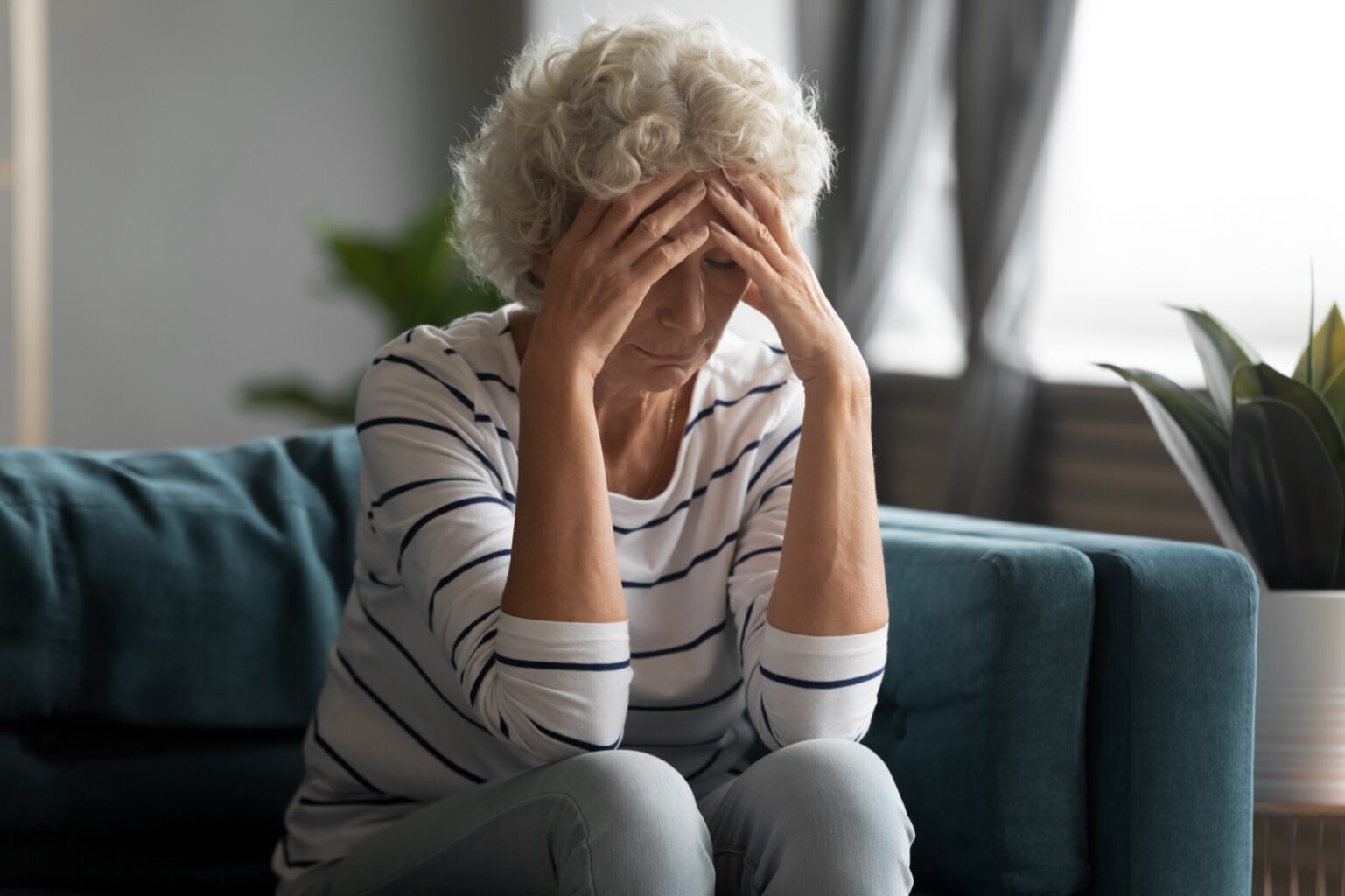 Unhealthy old senior retired woman sitting on sofa, suffering from strong headache. Unhappy elderly middle aged hoary grandmother having depressed mood, feeling insecure desperate lonely at home.